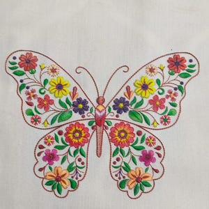 Op de afbeelding: Geborduurd vlinderontwerp met kleurrijke bloempatronen in rood, geel, paars en oranje op een crèmekleurige stof. De omtrek van de vlinder is bruin en de vleugels zijn gevuld met gedetailleerde bloem- en bladmotieven.
