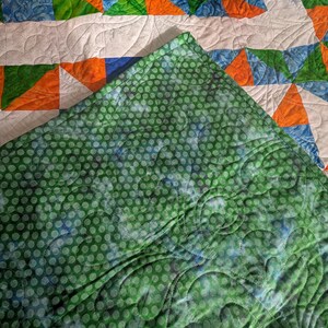 May include: A handmade quilt with a patchwork design. The quilt features orange, green, and blue triangles and curved shapes on a white background. The underside is green with a polka dot pattern.