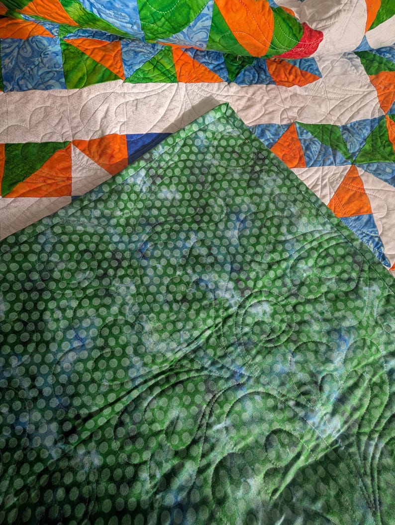 May include: A handmade quilt with a patchwork design. The quilt features orange, green, and blue triangles and curved shapes on a white background. The underside is green with a polka dot pattern.