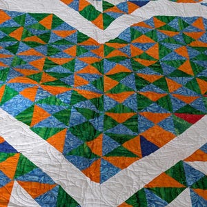 May include: A handmade quilt featuring a geometric design of triangles in shades of orange, green, and blue, set against a white background. The quilt has a heart-shaped pattern with white borders and quilted details.