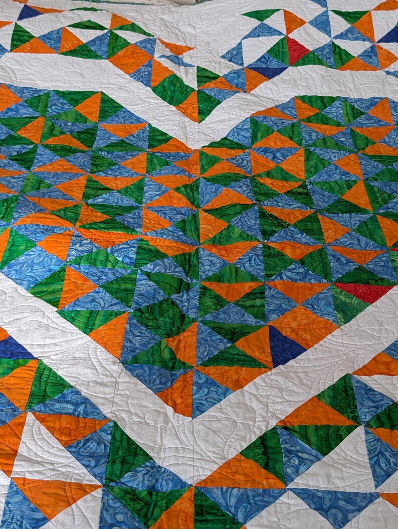 May include: A handmade quilt featuring a geometric design of triangles in shades of orange, green, and blue, set against a white background. The quilt has a heart-shaped pattern with white borders and quilted details.