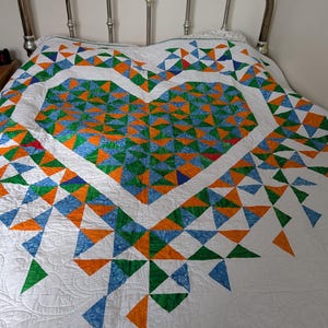 May include: A handmade quilt featuring a large heart design. The heart is composed of blue, orange, and green triangles on a white background. The quilt is on a bed with a silver metal headboard.