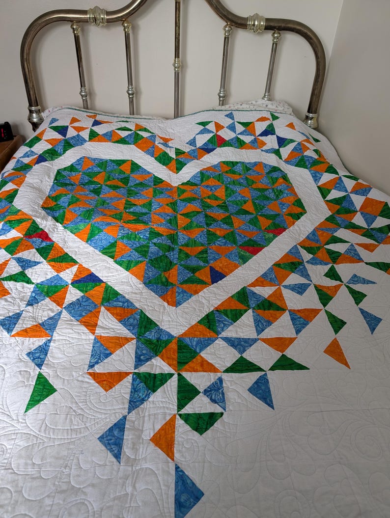 May include: A handmade quilt featuring a large heart design. The heart is composed of blue, orange, and green triangles on a white background. The quilt is on a bed with a silver metal headboard.