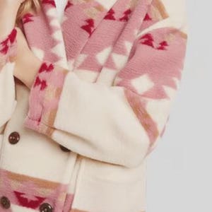 May include: Cream-colored coat with a geometric pattern in shades of pink, red, and brown. The coat features a double-breasted design with dark brown buttons. The sleeves and lapels showcase the patterned fabric.