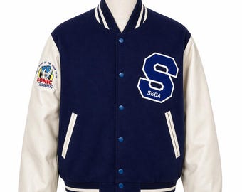 May include: A navy blue and white varsity jacket with the SEGA logo and a Sonic the Hedgehog patch. The jacket has white sleeves, a striped hem, and blue snap closures. The jacket is displayed on a mannequin.