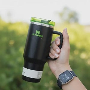 May include: Black Stanley insulated mug with a green lid and handle. The mug has the Stanley logo in green. The mug is held in a hand, and a smartwatch is visible on the wrist.