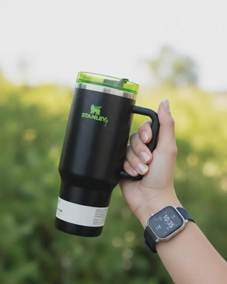 May include: Black Stanley insulated mug with a green lid and handle. The mug has the Stanley logo in green. The mug is held in a hand, and a smartwatch is visible on the wrist.