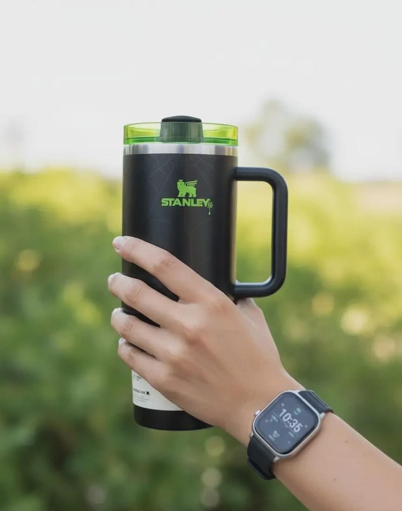 May include: Black Stanley mug with a green lid and handle. The mug has the Stanley logo in green. The mug is held in front of a blurred green background. A smartwatch is visible on the person's wrist.