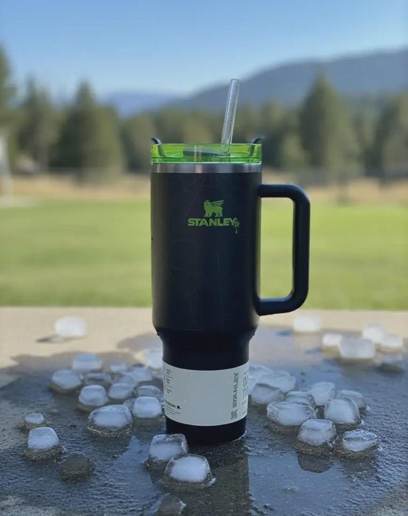 May include: Black Stanley travel mug with a green lid and handle, featuring the Stanley logo in green. A clear straw extends from the lid. The mug is surrounded by ice cubes on a wet surface, suggesting cold beverage use.