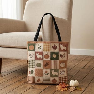 Könnte beinhalten: Eine rechteckige Tragetasche mit schwarzen Schulterriemen und einem Patchwork-Design aus Quadraten in Grün-, Braun- und Cremetönen. Das Muster zeigt Illustrationen von Kürbissen, Corgis, Herbstblättern, Blumen und Eicheln.