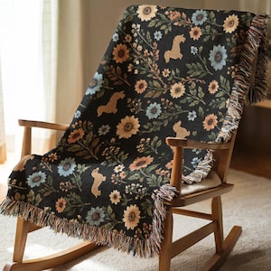 Peut inclure: Un plaid tissé noir orné d'un motif répétitif de tournesols jaunes, de fleurs bleu clair, de feuilles vertes et de silhouettes de lapins couleur beige. La couverture présente une finition texturée et des bords effilochés sur les côtés.