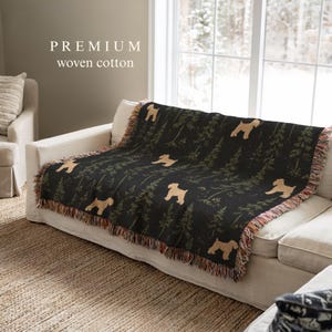 Op de afbeelding: Een geweven katoenen plaid met een herhalend patroon van beige hondensilhouetten en donkergroene dennenbomen op een zwarte achtergrond. De deken heeft een gefranjerde rand en de tekst PREMIUM woven cotton is zichtbaar in de linkerbovenhoek.