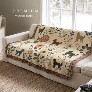 Peut inclure: Un plaid en coton tissé de couleur crème orné d'un motif répétitif de silhouettes de chiens, de grandes fleurs orange et bleues, et de feuillages verts. Le plaid présente des bords à franges et le texte PREMIUM woven cotton imprimé au-dessus.