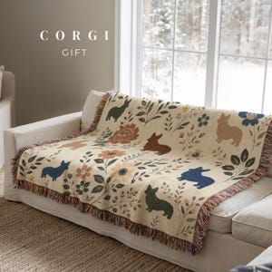 May include: A cream-colored woven throw blanket featuring a repeating pattern of Corgi dogs in blue, brown, and green, interspersed with various floral and botanical motifs in muted tones. The blanket has a fringed edge and text reading CORGI GIFT.