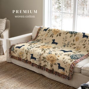 Peut inclure: Un plaid en coton tissé présentant un fond crème avec un motif répétitif de silhouettes de teckels bleu foncé, de roses jaunes et de feuillage vert. La couverture possède un bord à franges et le texte PREMIUM woven cotton.