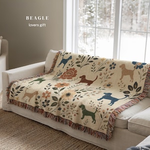 Puede incluir: Una manta tejida de color crema con un patrón repetitivo de beagles en varios colores, incluyendo azul, marrón y verde, intercalados con motivos florales y botánicos. El texto BEAGLE lovers gift está impreso en la esquina superior izquierda.