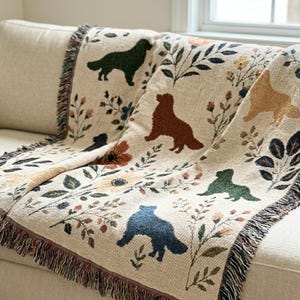 May include: A woven throw blanket featuring a cream-coloured background with a repeating pattern of silhouettes of dogs in various colours including dark green, rust brown, and navy blue, interspersed with colourful floral and botanical motifs.