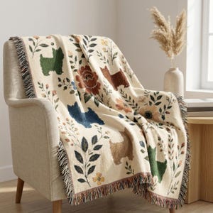 May include: A cream-coloured woven throw blanket featuring a repeating pattern of small dog silhouettes in green, blue, and brown, interspersed with colourful floral motifs and leafy branches. The blanket has a fringed edge along the sides.