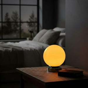 May include: A spherical table lamp with a glowing yellow glass globe shade and a metallic base. The lamp emits a warm, soft light, illuminating the immediate area around it. The design is simple, featuring a smooth, round glass orb.