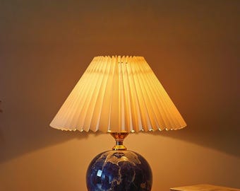 May include: A table lamp featuring a pleated, off-white conical lampshade and a spherical base with a deep blue and white marbled pattern. The lamp includes a gold-toned metal neck connecting the base to the shade, emitting a warm light.