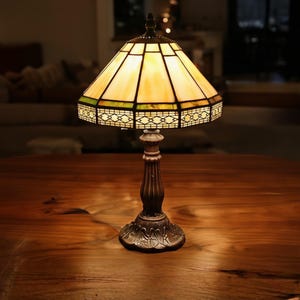 May include: A table lamp featuring a stained glass shade with cream-colored panels and a green geometric border. The lamp has a dark bronze-toned metal base with a fluted column and an ornate, leaf-patterned circular foot, providing warm illumination.
