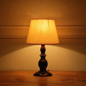 Może przedstawiać: Ciemnobrązowa lampa stołowa z beżowym abażurem. Klosz ma kształt stożka, a podstawa ma dekoracyjny, toczony wzór. Lampa stoi na drewnianej powierzchni, rzucając ciepłą poświatę.