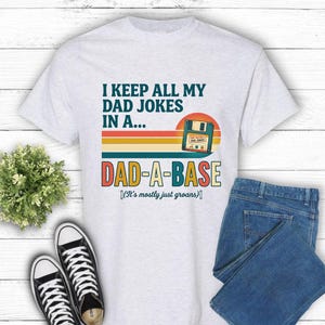 Könnte beinhalten: Ein hellgraues T-Shirt mit dem Text "I KEEP ALL MY DAD JOKES IN A... DAD-A-BASE (It's mostly just groans)". Das Shirt wird mit einer blauen Jeans und schwarz-weißen Turnschuhen kombiniert.
