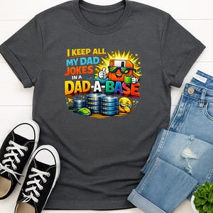 Könnte beinhalten: Dunkelgraues T-Shirt mit dem Text "I KEEP ALL MY DAD JOKES IN A DAD-A-BASE" und einer bunten Grafik einer Computerdiskette mit Sonnenbrille, einer Kaffeetasse und Datenbank-Symbolen. Schwarze Turnschuhe und blaue Jeans sind ebenfalls im Bild.