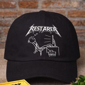May include: Black baseball cap with the text "FUNNY HAT" in yellow and a white graphic of a person sleeping at a computer with the text "REST AREA". The hat has a curved brim and a "BEST SELLER" tag.