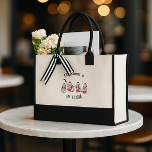 May include: A canvas tote bag with black handles and a black bottom panel. The bag features the text "Christmas is so fetch!" and an illustration of four women wearing Santa hats. A bouquet of flowers and a striped ribbon adorn the bag.