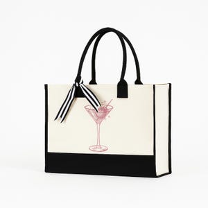 May include: A canvas tote bag with a black base and handles. The bag features a pink illustration of a cocktail in a martini glass. A black and white striped ribbon is tied to one of the handles. The bag is on a white background.