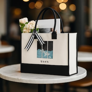 May include: A cream-colored tote bag with black accents and handles, featuring a Japanese-style wave design. The bag is filled with white flowers and a striped ribbon. The bag is sitting on a marble table.