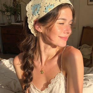 Può includere: Una fascia per capelli all'uncinetto color crema con dettagli di margherite blu. Sono visibili anche una collana con pendente dorato e una camisole bianca con stampa floreale.