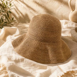 Peut inclure: Chapeau de soleil tissé de couleur marron clair avec un large bord incurvé. Le chapeau est fait de fibres naturelles et repose sur un tissu crème. L'image montre des ombres projetées sur le tissu et le chapeau.