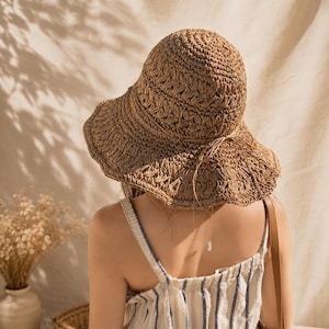 May include: A woven straw sun hat with a wide, wavy brim. The hat is a light brown color and has a textured pattern. The hat is worn with a striped tank top and a brown shoulder bag. The background is a neutral beige.
