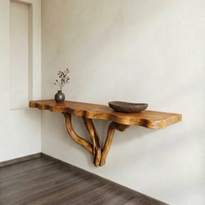 Op de afbeelding: Een houten console met een live edge design. De tafel wordt ondersteund door drie takachtige poten en is aan een witte muur bevestigd. Een kleine vaas met gedroogde bloemen en een houten kom staan op de tafel. De tafel is ongeveer 152 cm lang.