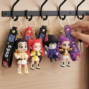 Może przedstawiać: Kolekcja breloków z postaciami K-pop zawieszonych na czarnym metalowym wieszaku. Każdy brelok przedstawia miniaturową figurkę z unikalnymi strojami i fryzurami. Breloki mają pasek z napisem "K-pop".