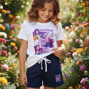 May include: A young person wearing a white t-shirt and navy blue shorts. The t-shirt features a colorful graphic with the text "HUNTRA RUM MIRA ZOR". The shorts have a white drawstring and a small graphic on the leg.