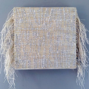 May include: A beige rectangular wall hanging made of woven strips of paper with frayed edges. The paper is covered in small, white printed text.