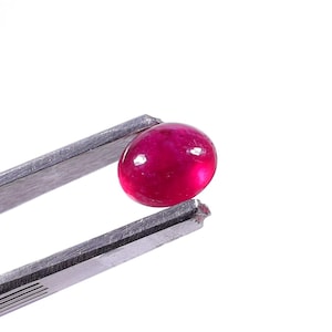 Könnte beinhalten: Ein ovaler Rubin-Edelstein, gehalten von einer silbernen Pinzette. Der Rubin hat eine tiefrote Farbe mit polierter, glänzender Oberfläche. Die Pinzette besteht aus glänzendem Metall, vor weißem Hintergrund.