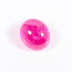 Puede incluir: Una gema pulida, de forma ovalada, en un tono magenta brillante. La piedra tiene una superficie lisa y brillante con sutiles variaciones internas y una ligera translucidez. La gema se encuentra sobre un fondo blanco.