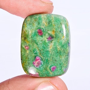Puede incluir: Una gema rectangular pulida con esquinas redondeadas. La piedra presenta un vibrante color base verde con inclusiones rojas y rosas dispersas. La superficie tiene un acabado brillante, resaltando los patrones y texturas naturales dentro de la piedra.