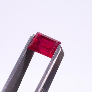 Puede incluir: Un rubí de corte rectangular, sujeto con unas pinzas metálicas. La gema de color rojo intenso se encuentra sobre un fondo blanco liso. Las facetas del rubí capturan la luz, resaltando su claridad y brillo.