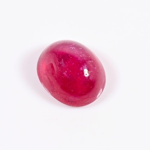 May include: A polished, oval-shaped ruby gemstone. The gem is a deep, saturated red colour with a smooth, glossy surface. The ruby is set against a plain white background, highlighting its colour and shape.