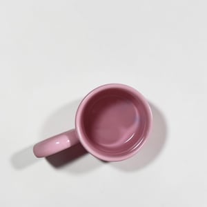May include: A small, pink ceramic mug with a handle. The mug is a solid, soft pink color and appears to be empty. The mug is set against a white background.