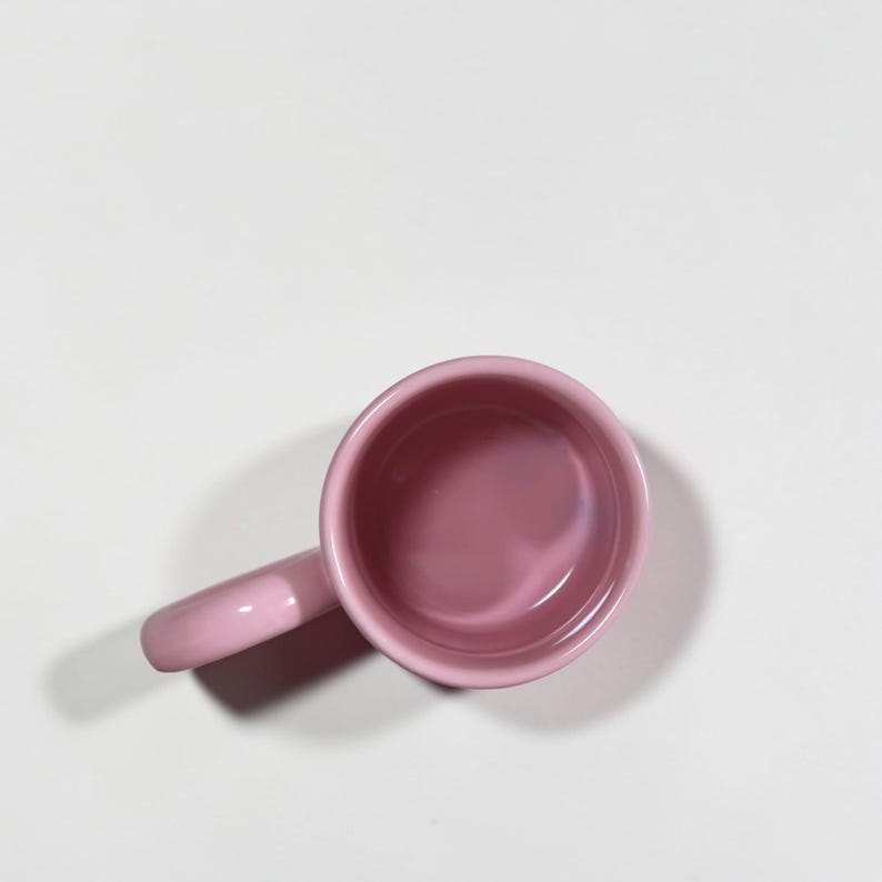 May include: A small, pink ceramic mug with a handle. The mug is a solid, soft pink color and appears to be empty. The mug is set against a white background.