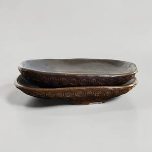 Może przedstawiać: Dwa owalne ceramiczne naczynia ułożone jedno na drugim. Górne naczynie jest w stonowanym szaro-niebieskim kolorze, a dolne w ciemniejszym brązie. Oba mają teksturowany, spiralny wzór na obrzeżu, sugerujący rustykalną lub ręcznie robioną estetykę. Wymiary w cm.