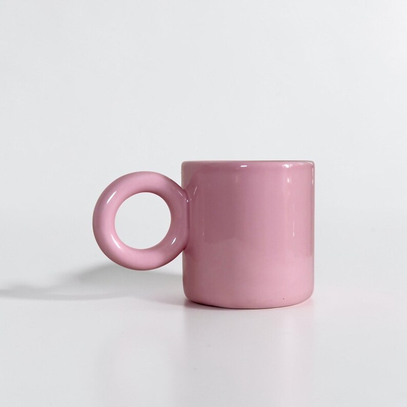 May include: A pink ceramic mug with a unique circular handle. The mug is cylindrical in shape and has a smooth, glossy finish. The handle is a large, rounded loop, providing a comfortable grip. The mug is set against a plain white background.
