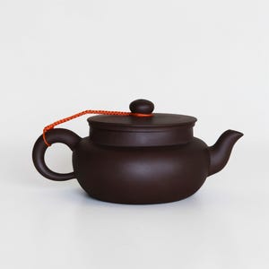 Może przedstawiać: Ciemnobrązowy czajnik z pasującą pokrywką i zakrzywionym uchwytem. Pomarańczowy sznurek jest przymocowany do uchwytu i pokrywki. Czajnik ma zaokrąglony korpus i dzióbek.