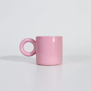 May include: A solid pink ceramic mug with a rounded handle. The mug has a cylindrical body and a smooth, glossy finish. The handle is a large, circular loop. The mug is set against a white background.